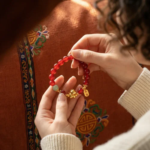 Hands holding a 2026 Year of the Fire Horse feng shui bracelet made of natural red agate beads, gold horse charm and jade accent bead, against a warm terracotta Tibetan robe embroidered with traditional dragon and lotus motifs and a Tibetan decorative border