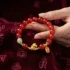 Natural red agate feng shui bracelet for 2026 Year of the Fire Horse, featuring a gold horse zodiac charm and 平安 (Peace) blessing tag, held by hand over a deep burgundy Tibetan brocade fabric embroidered with Om mantra symbols