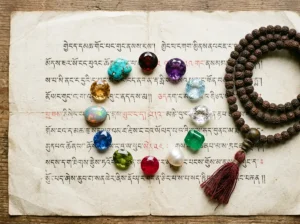A flat lay arrangement of 12 gemstones representing each birth month, including garnet, amethyst, aquamarine, diamond, emerald, pearl, ruby, peridot, sapphire, opal, topaz, and turquoise, placed on aged Tibetan manuscript paper with soft natural light