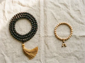 Flat lay product photography, top-down view. A full 108-bead mala necklace made of dark bodhi seeds with a golden tassel, arranged in a circular loop on the left side. On the right side, a 27-bead wrist mala bracelet made of light sandalwood beads with a small guru bead. Both items on a textured natural linen cloth with soft warm morning light from the upper left. Minimal, clean composition with generous negative space between items. Soft shadows. No text. Lifestyle spiritual aesthetic. Shot on medium format camera, f/4.0, warm neutral tones.
