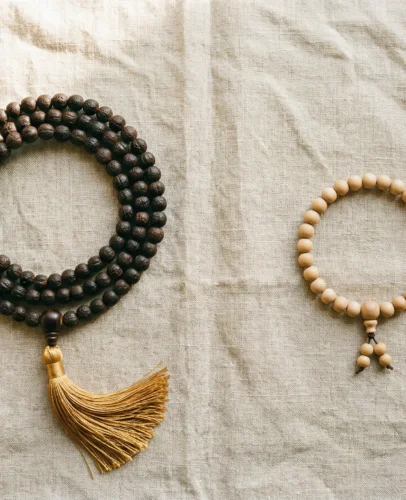 Flat lay product photography, top-down view. A full 108-bead mala necklace made of dark bodhi seeds with a golden tassel, arranged in a circular loop on the left side. On the right side, a 27-bead wrist mala bracelet made of light sandalwood beads with a small guru bead. Both items on a textured natural linen cloth with soft warm morning light from the upper left. Minimal, clean composition with generous negative space between items. Soft shadows. No text. Lifestyle spiritual aesthetic. Shot on medium format camera, f/4.0, warm neutral tones.