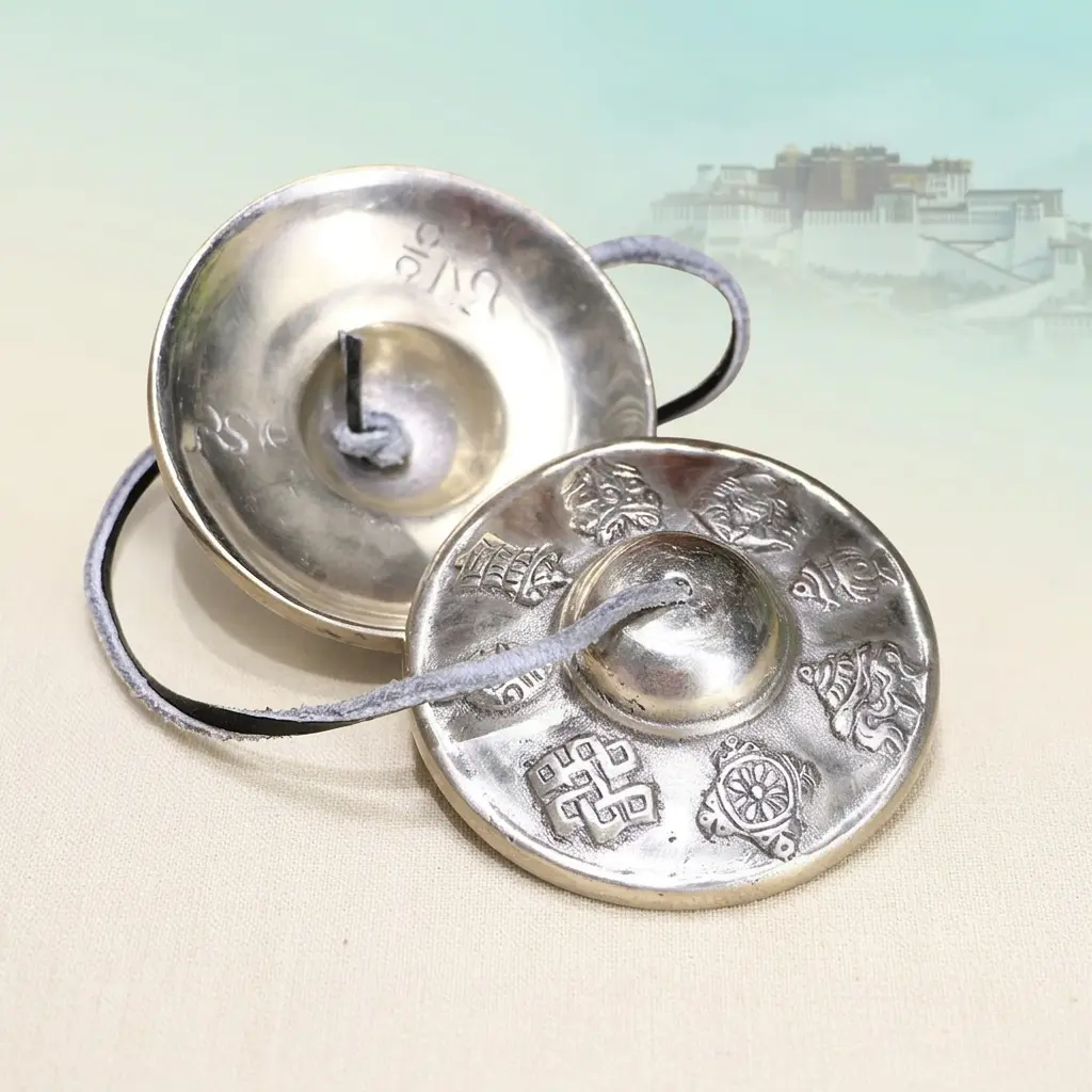 tibetan-tingsha-bells-interior-exterior-potala-palace-potalastore Two white copper Tibetan tingsha bells showing engraved Eight Auspicious Symbols on the outer face and polished concave interior, with Potala Palace in the background