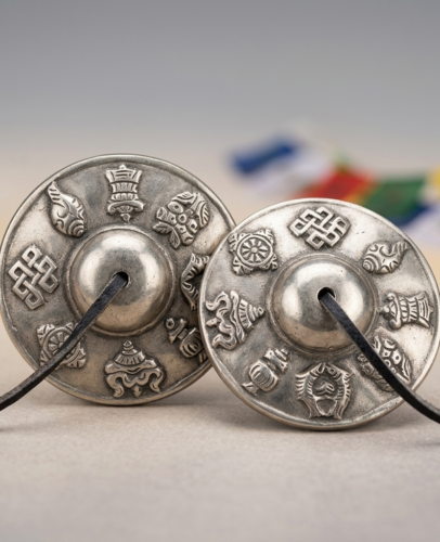 A matched pair of white copper Tibetan tingsha bells showing Ashtamangala engravings, with Tibetan prayer flags blurred in the background