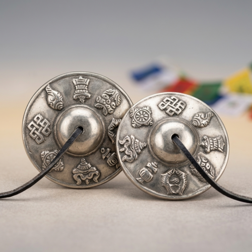 A matched pair of white copper Tibetan tingsha bells showing Ashtamangala engravings, with Tibetan prayer flags blurred in the background