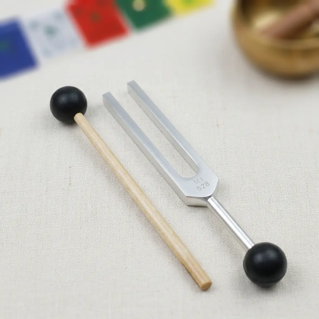 528 Hz Tuning Fork and Mallet Set with Tibetan Prayer Flag Background – PotalaStore Sound Healing 528 Hz aluminum alloy tuning fork and wood-handle mallet laid flat on linen, with colorful Tibetan prayer flags and a singing bowl in the background — PotalaStore