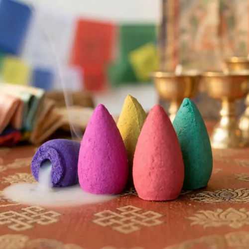 Four colorful cone incenses in purple, pink, yellow and turquoise with white smoke rising, arranged on Tibetan altar cloth with prayer flags and brass bowls