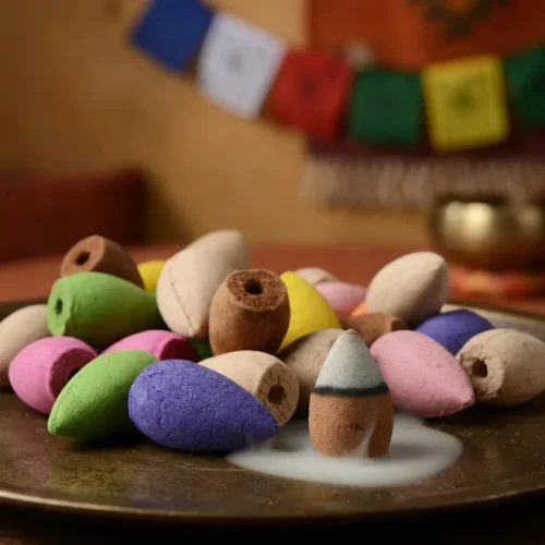 Assorted colorful backflow incense cones in pink, yellow, green, purple, beige and brown arranged on brass plate with Tibetan prayer flags in background, natural product photography