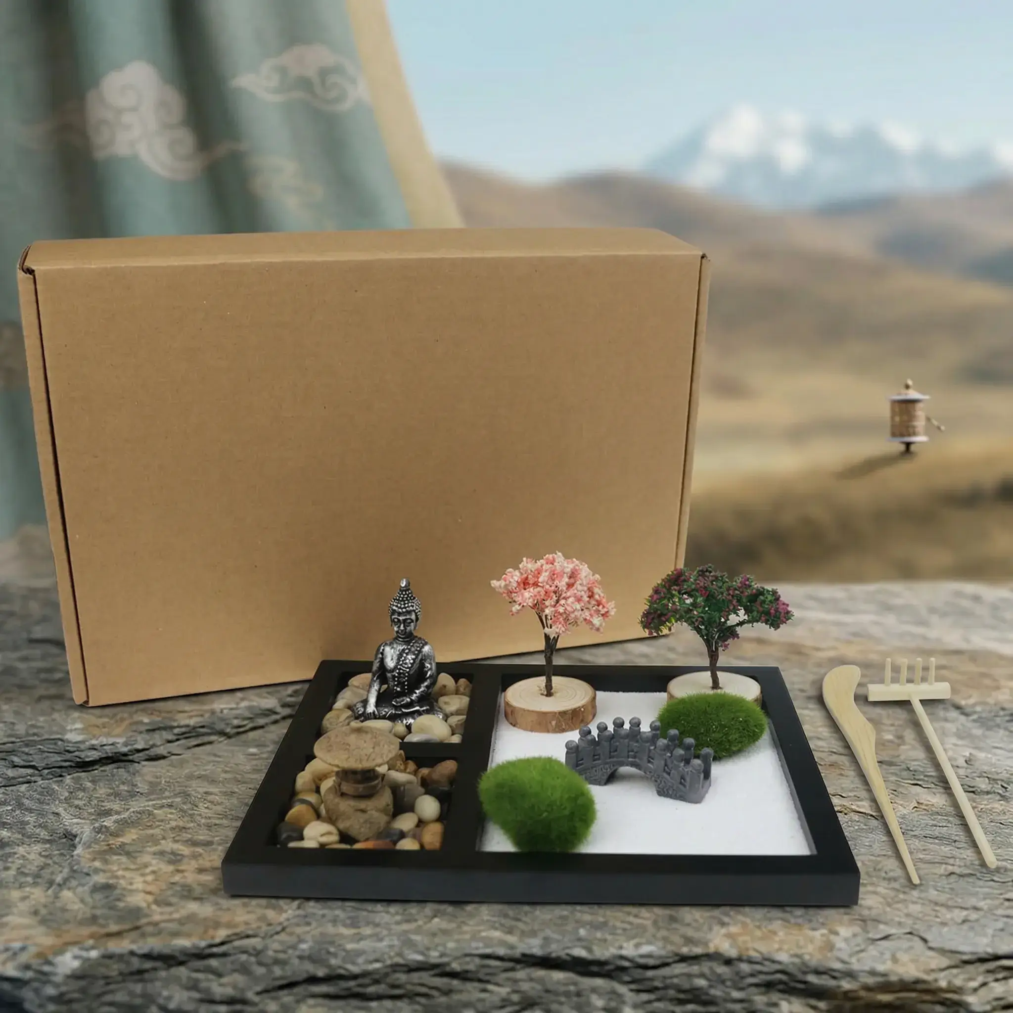 Complete Buddha Zen Garden Kit with Box and Bamboo Rake Tools – Tibetan Highland Full Buddha zen garden desk kit showing meditating Buddha statue, miniature cherry blossom and flowering trees, stone arch bridge, moss balls, natural rocks, sand, bamboo rake tools, and kraft gift box, photographed on Tibetan highland rocks with a prayer wheel visible in the distance — PotalaStore