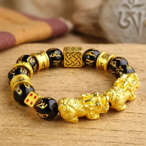 Close-up side view of double Pixiu bracelet showing detailed gold-plated Pixiu amulet with dragon head, red dot lucky dice charm, and black obsidian mantra beads against a textured wooden background
