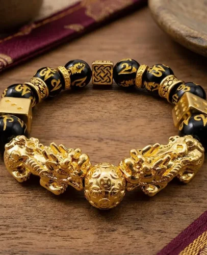 Double Pixiu Feng Shui Wealth Bracelet – Black Obsidian with Lucky Dice Charms Double Pixiu feng shui bracelet flat lay on wooden surface showing gold wealth ball centerpiece, two gold lucky dice charms, Celtic knot bead, and Om Mani Padme Hum engraved obsidian beads