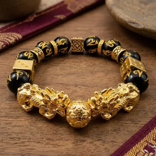 Double Pixiu feng shui bracelet flat lay on wooden surface showing gold wealth ball centerpiece, two gold lucky dice charms, Celtic knot bead, and Om Mani Padme Hum engraved obsidian beads