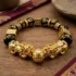 Double Pixiu feng shui bracelet flat lay on wooden surface showing gold wealth ball centerpiece, two gold lucky dice charms, Celtic knot bead, and Om Mani Padme Hum engraved obsidian beads