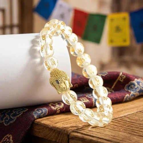 Gold Buddha mantra bracelet featuring 8mm clear crystal beads engraved with Om Mani Padme Hum and a gold micro-pavé Buddha head, styled with colorful Tibetan prayer flags and monastery brocade
