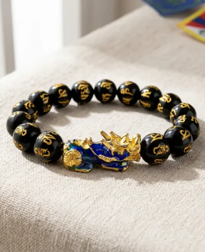 Tibetan Buddhist feng shui black obsidian Pixiu bracelet with gold-painted mantra beads and cloisonné Pixiu charm, resting on linen surface with Tibetan flag in background