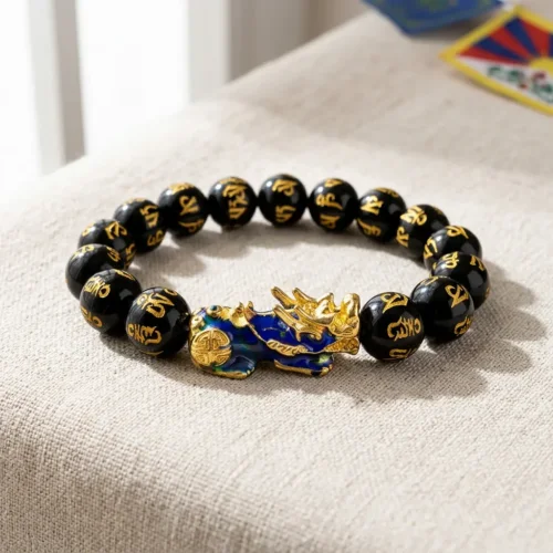 Tibetan Buddhist feng shui black obsidian Pixiu bracelet with gold-painted mantra beads and cloisonné Pixiu charm, resting on linen surface with Tibetan flag in background