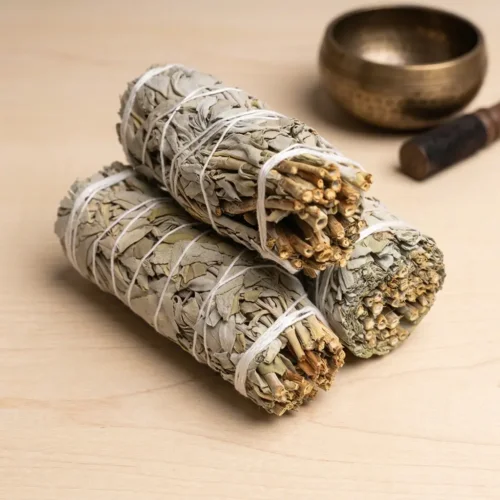 Five California white sage smudge sticks stacked on a stone surface next to a copper Tibetan prayer wheel, ideal for home cleansing and smudging rituals