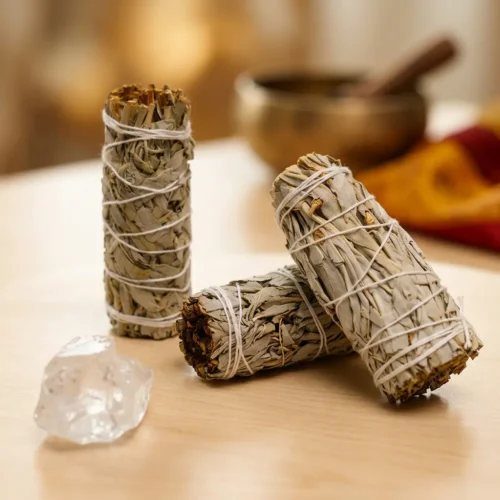 Three upright white sage smudge sticks displayed with a clear quartz crystal and Tibetan singing bowl, surrounded by colorful Tibetan textile, for meditation and space clearing