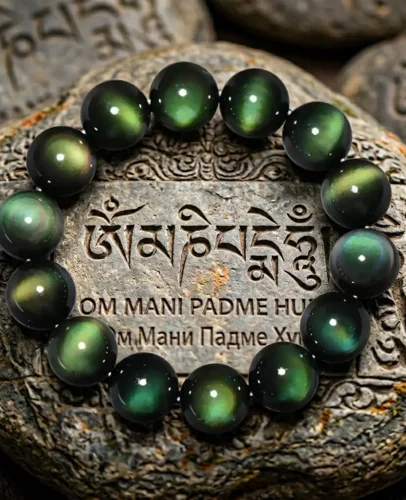Green eye obsidian healing bracelet placed on carved Tibetan mani stone engraved with Om Mani Padme Hum mantra in Sanskrit and Tibetan script, rainbow obsidian beads glowing with green and gold iridescence