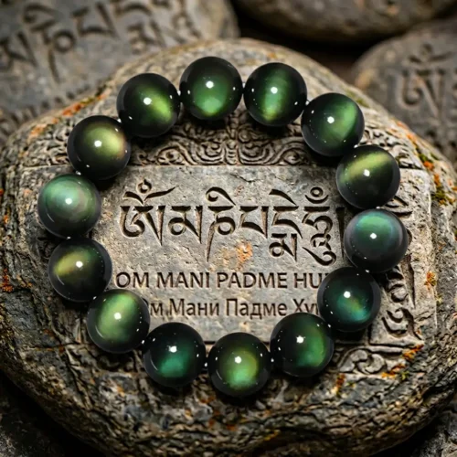 Green eye obsidian healing bracelet placed on carved Tibetan mani stone engraved with Om Mani Padme Hum mantra in Sanskrit and Tibetan script, rainbow obsidian beads glowing with green and gold iridescence