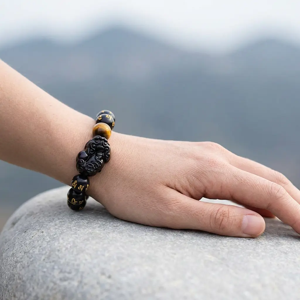obsidian-pixiu-bracelet-worn-on-left-wrist-outdoor Person wearing black obsidian Pixiu wealth bracelet on left wrist resting on stone, mountain landscape in background