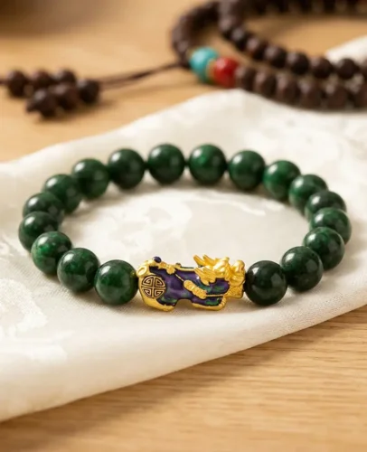 piyao-jade-bracelet-flatlay-cloisonne-pixiu-shou-symbol Close-up flat lay of a green jade Piyao bracelet on white silk cloth, showing the gold cloisonné Pixiu charm engraved with a Shou longevity symbol, with a Tibetan mala in the background