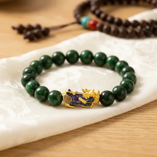 Close-up flat lay of a green jade Piyao bracelet on white silk cloth, showing the gold cloisonné Pixiu charm engraved with a Shou longevity symbol, with a Tibetan mala in the background