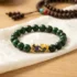 Close-up flat lay of a green jade Piyao bracelet on white silk cloth, showing the gold cloisonné Pixiu charm engraved with a Shou longevity symbol, with a Tibetan mala in the background