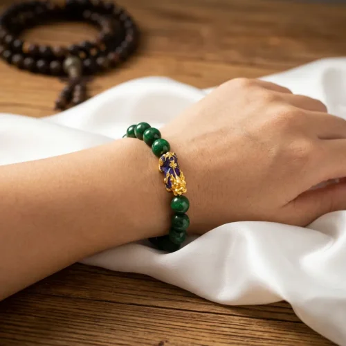 Woman wearing a 12mm green jade Piyao bracelet on her left wrist, featuring a gold and purple cloisonné Pixiu charm, styled with a Tibetan Buddhist mala in the background