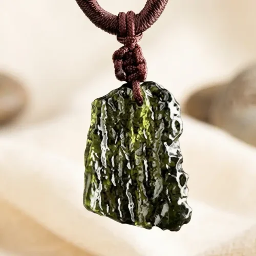 Close-up of raw moldavite green tektite pendant hanging on dark red braided cord against soft beige zen stone background