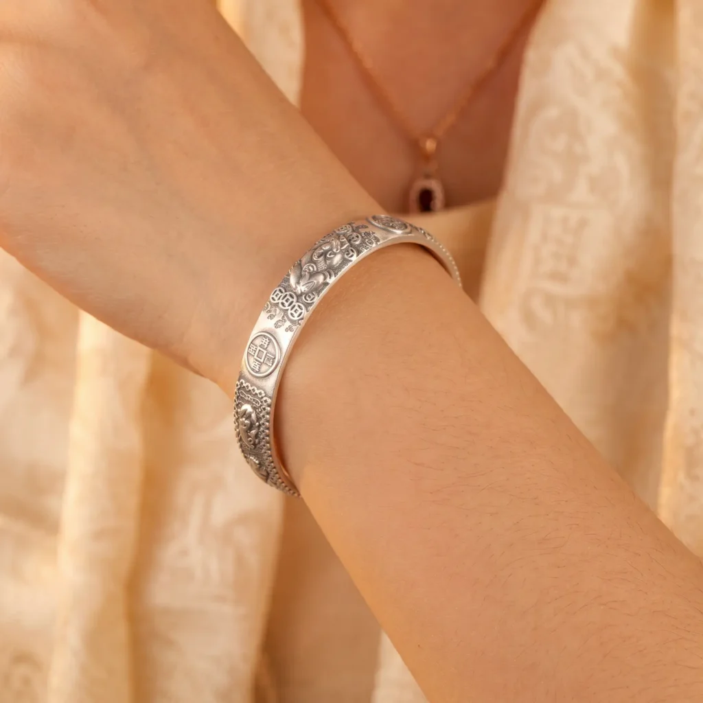 Braccialetto portafortuna Fortune Pixiu in argento, indossato al polso su sfondo di seta tibetana dorata. Bracciale portafortuna Pixiu in argento al polso, su tessuto di seta tibetana ricamato in oro, che mostra in dettaglio le incisioni della bestia della ricchezza Pixiu e delle monete portafortuna.