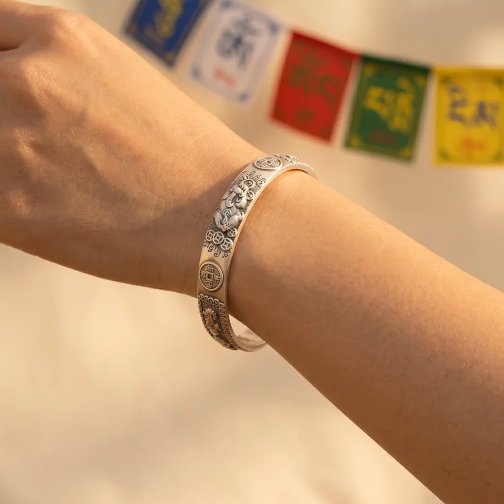 Braccialetto portafortuna Pixiu indossato al polso sinistro con bandiere di preghiera tibetane. Bracciale portafortuna Pixiu indossato al polso sinistro con colorate bandiere di preghiera tibetane sullo sfondo, che mostrano la bestia Pixiu e incisioni di monete portafortuna cinesi.