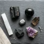 best-crystals-protection-negative-energy-flatlay Seven best crystals for protection from negative energy arranged on a dark surface, including black tourmaline, black obsidian, hematite, amethyst, and selenite.