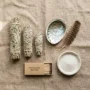 Smudging Essentials_ White Sage, Abalone Shell & More A flat-lay of white sage smudge sticks, an abalone shell, a feather, matches, and a small dish of sand arranged on a neutral linen surface