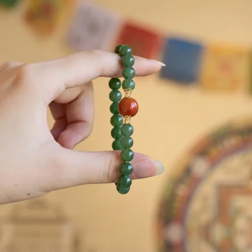 Hand holding a Hetian Jasper & Red Agate Luck Bracelet with a gold-capped Nanhong agate bead, Tibetan prayer flags behind
