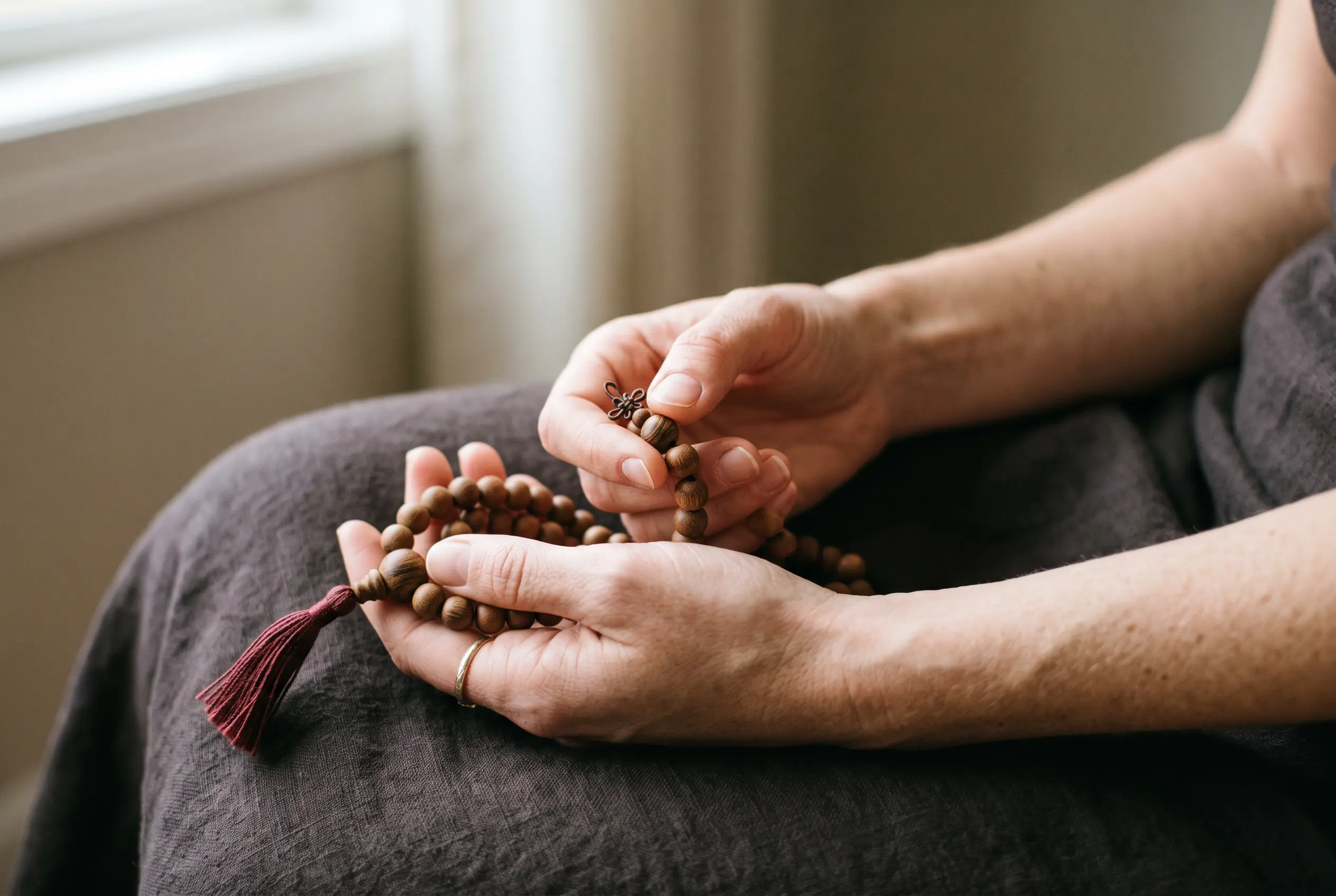 Hands holding a sandalwood mala, thumb moving one bead during japa meditation.