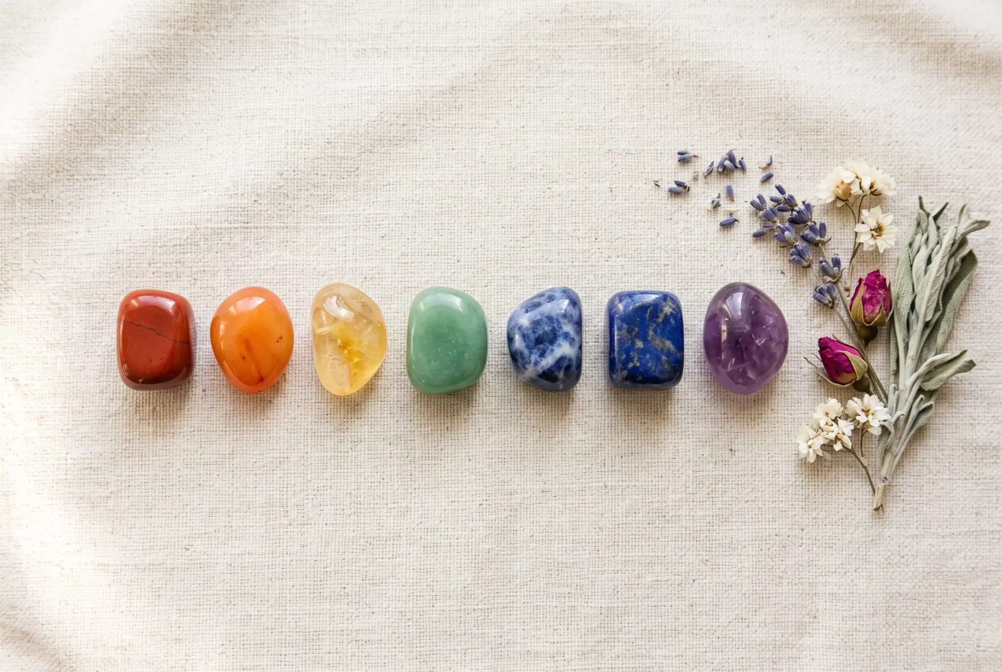 Seven tumbled healing crystals arranged in a row in chakra colors from red to violet on a natural linen background