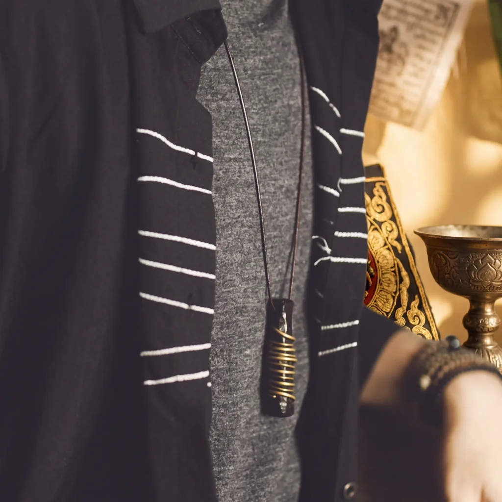Ebony wood and brass pendant necklace worn over a grey tee and black jacket, with a Tibetan prayer flag and carved brass cup behind