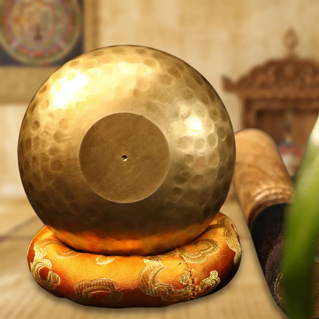 base a ciotola tibetana con vista, disco liscio e corpo martellato Parte inferiore di una ciotola tibetana in rame martellato con un disco di base liscio e piatto, poggiante su un cuscino di broccato arancione.