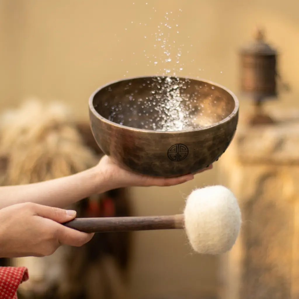 singing-bowl-water-sound-ritual-felt-mallet Hands holding a Tibetan singing bowl with water droplets splashing inside while a white felt mallet is held beside it