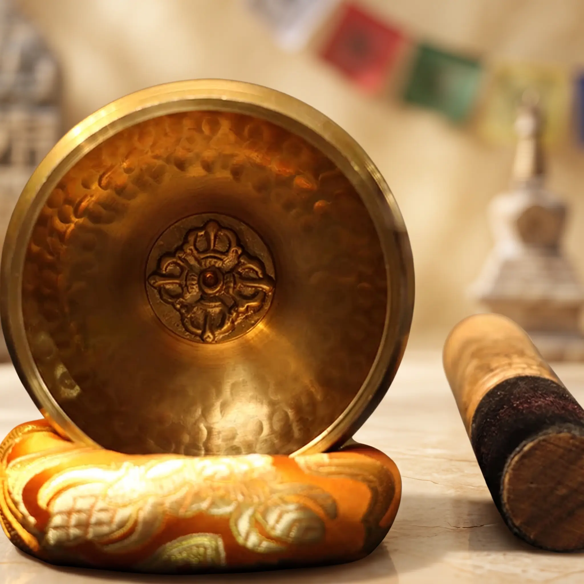 bandiere di preghiera con doppio vajra e ciotola tibetana Ciotola tibetana in rame con un emblema a doppio vajra fuso al centro, su un cuscino arancione accanto a un battente in legno.