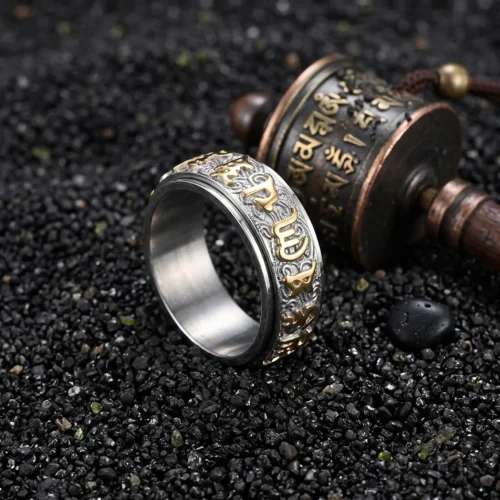 Cincin roda doa Tibet dua warna dengan aksara mantra emas di samping roda doa di atas pasir vulkanik hitam.